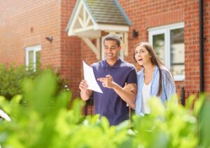 A couple standing outside a UK house, smiling and reviewing property details during their first home viewing