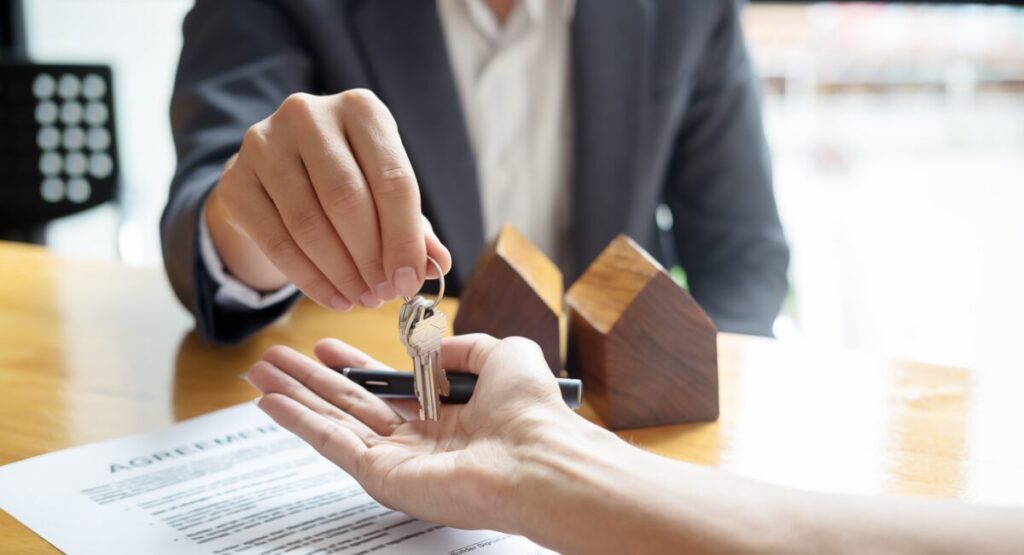 Close-up of landlord handing keys to a tenant over a rental agreement with wooden house models on the desk