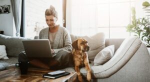 A person sitting on a sofa with a laptop and a dog beside them, representing a landlord managing a rental property from home.