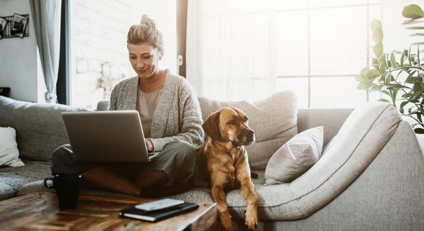 A person sitting on a sofa with a laptop and a dog beside them, representing a landlord managing a rental property from home.