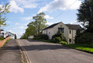 A quiet road with double yellow lines rising over a canal bridge with a white parapet, lined by a red brick wall on the left and a cream-rendered cottage