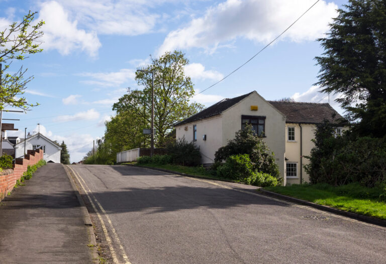 A quiet road with double yellow lines rising over a canal bridge with a white parapet, lined by a red brick wall on the left and a cream-rendered cottage