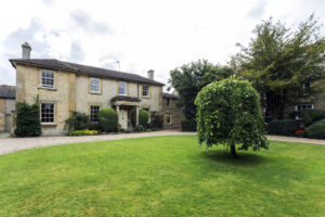 Two-storey honey-coloured stone house with white sash windows, a columned porch, and rounded topiary shrubs by the front door, set behind a gravel drive and a large green lawn with a small weeping tree.
