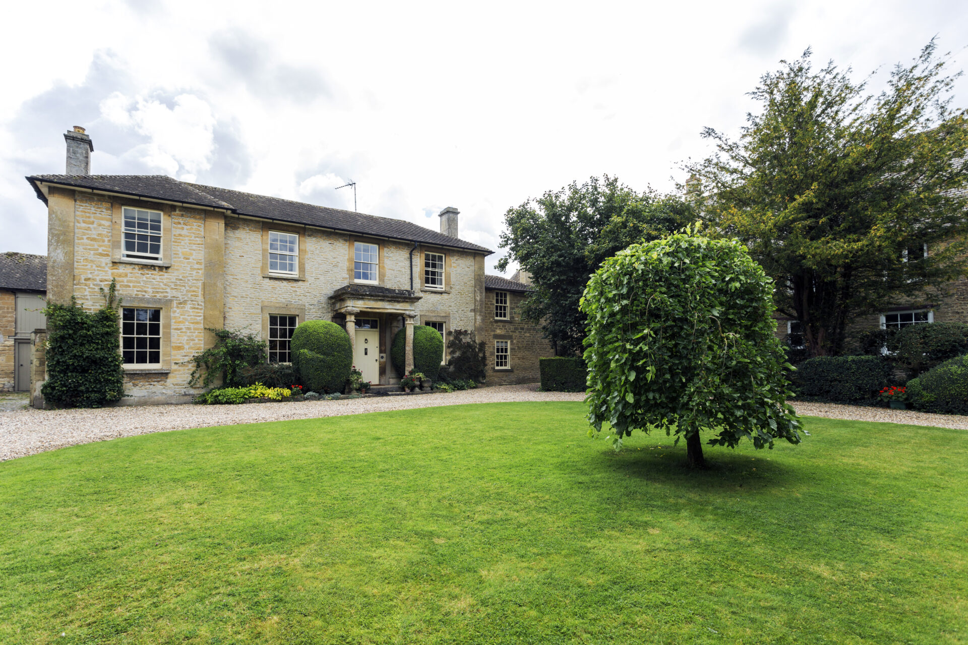 Two-storey honey-coloured stone house with white sash windows, a columned porch, and rounded topiary shrubs by the front door, set behind a gravel drive and a large green lawn with a small weeping tree.